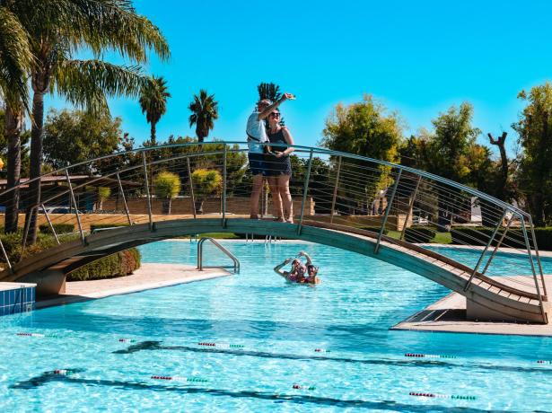 Coppia sul ponte, persone in piscina, giornata soleggiata.