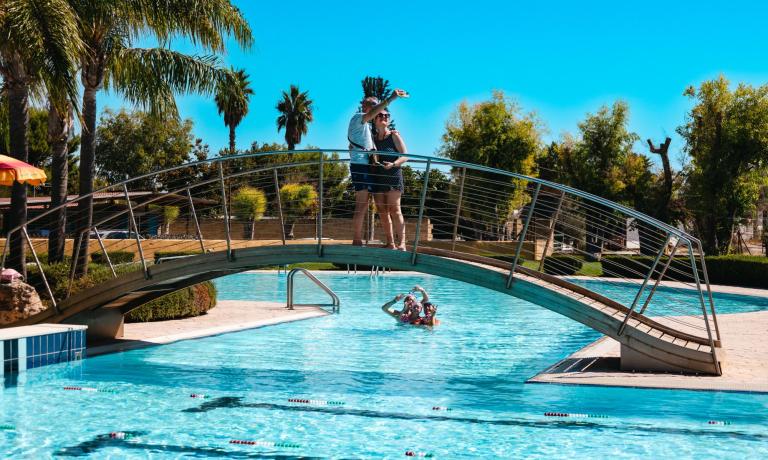 Coppia sul ponte, persone in piscina, giornata soleggiata.